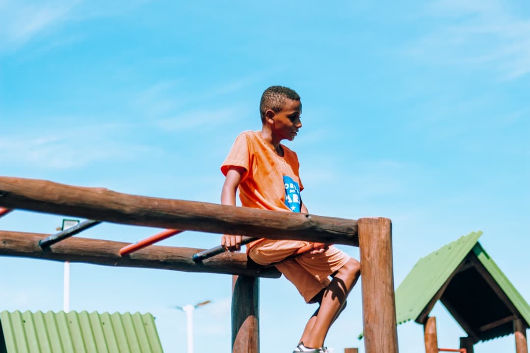 Um menino negro brincando em um parquinho de madeira ao ar livre, sob um céu azul claro e ensolarado. Ele está sentado em uma estrutura de troncos, com as pernas de um lado e o tronco do outro, em uma pose que sugere movimento e diversão. O menino veste uma camiseta laranja e bermuda da mesma cor, com um tênis esportivo. Na camiseta há a logo do instituto em azul, em formato de bandeira de São João, contendo o texto 'CORES DO MARÁ' e uma estrela. Ao fundo, aparecem telhados verdes de outras estruturas do parquinho. O formato da imagem tem cantos arredondados e bandeirinhas coloridas nos cantos — reforçando um tom alegre e infantil.