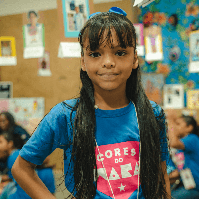 Menina sorrindo para a câmera em sala de aula, usando camiseta azul, com outras crianças e atividades ao fundo.